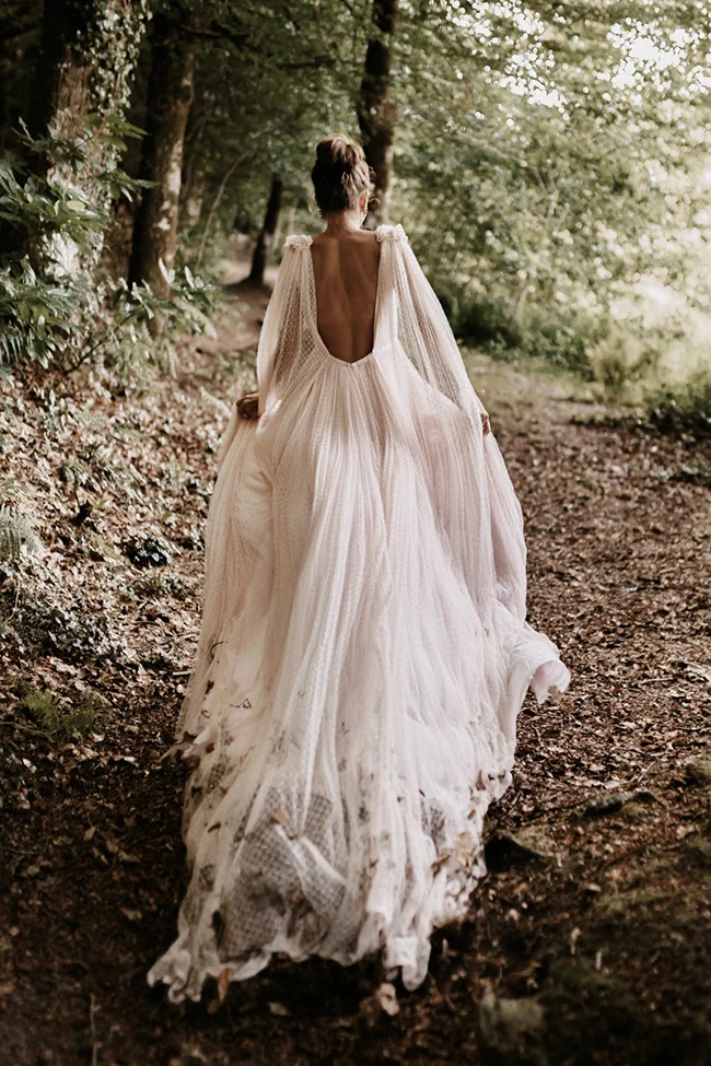 Mariée de dos en robe fluide et cape vaporeuse, marchant dans une forêt aux tons naturels et romantiques.