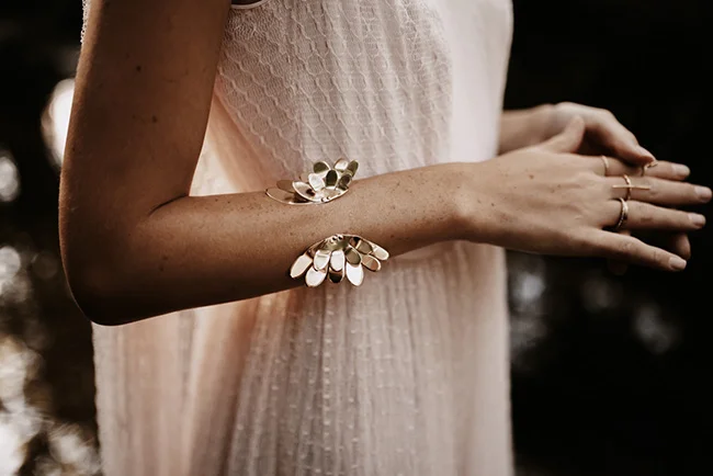 Gros plan d'une mariée en robe blanche portant des bracelets et bagues dorés sculpturaux dans une ambiance naturelle.