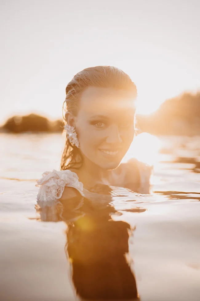 Mariée glamour souriante dans l'eau au coucher du soleil, portant une robe blanche et des bijoux, ambiance chaleureuse.