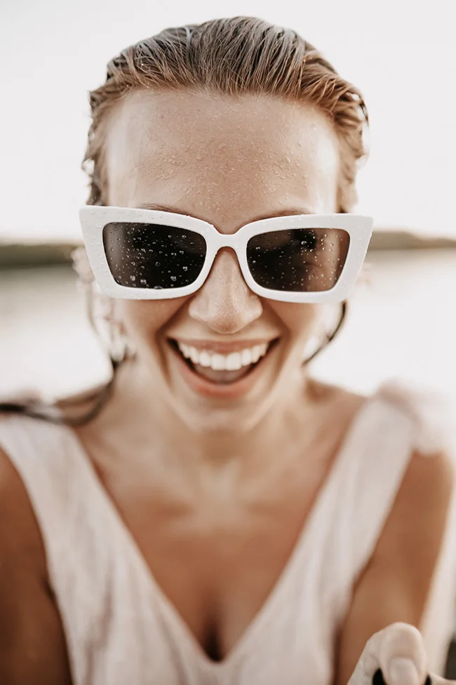 Une mariée souriante aux cheveux mouillés portant des lunettes de soleil blanches, dans une ambiance lumineuse et moderne.