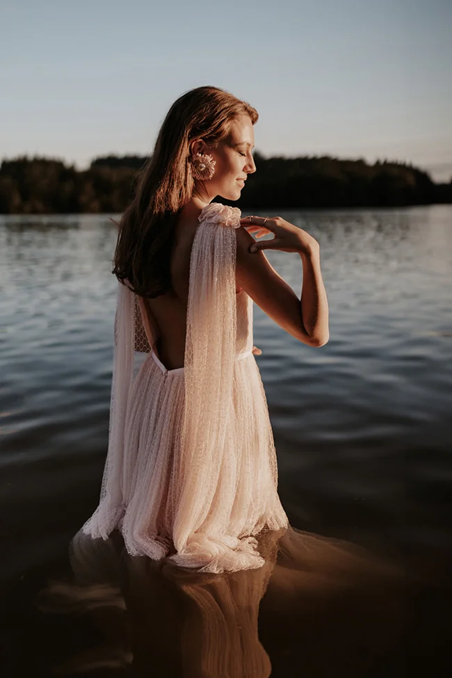 Mariée glamour en robe blanche vaporeuse, debout dans l'eau au coucher du soleil, ambiance romantique et sauvage.