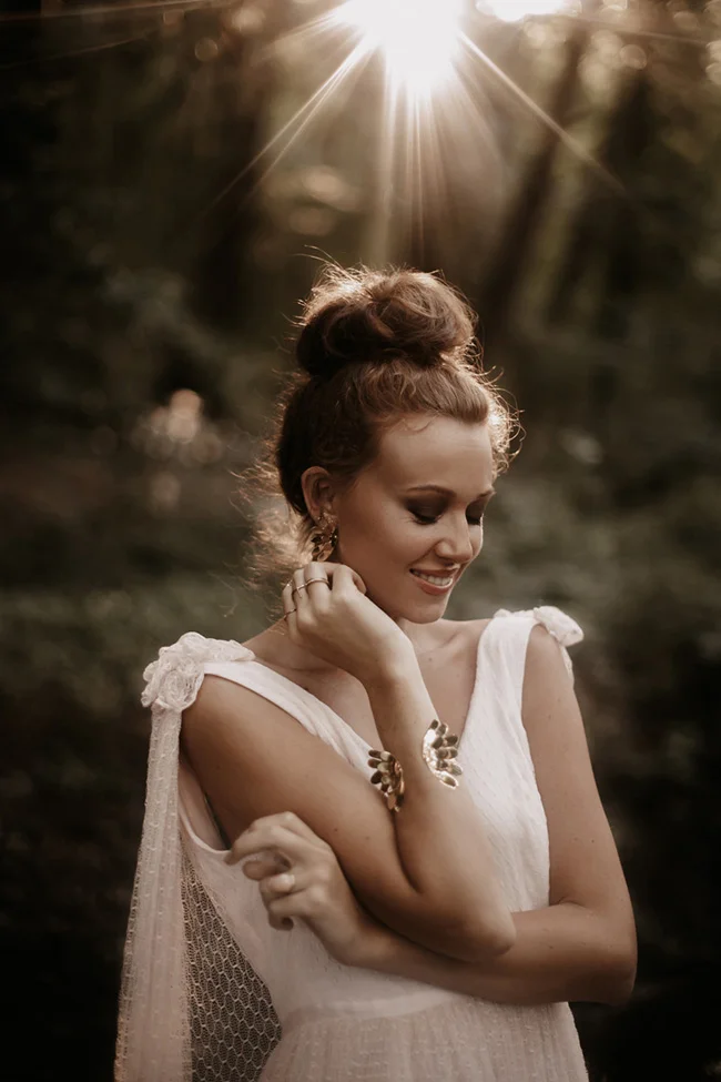 Mariée glamour en robe blanche et chignon, souriante sous un soleil éclatant dans un décor naturel et boisé.