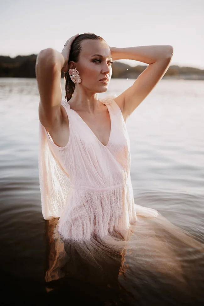 Une femme en robe rose pâle vaporeuse pose dans l'eau au coucher du soleil, dans une ambiance glamour et sauvage.