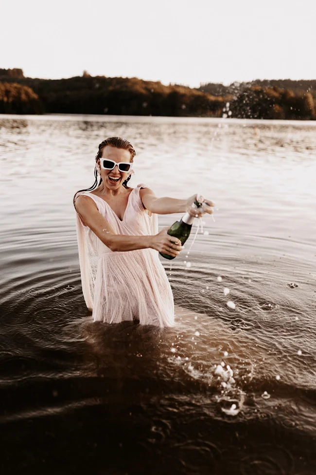 Une mariée en robe rose pâle et lunettes blanches sabrant le champagne dans un lac, ambiance festive et décalée.