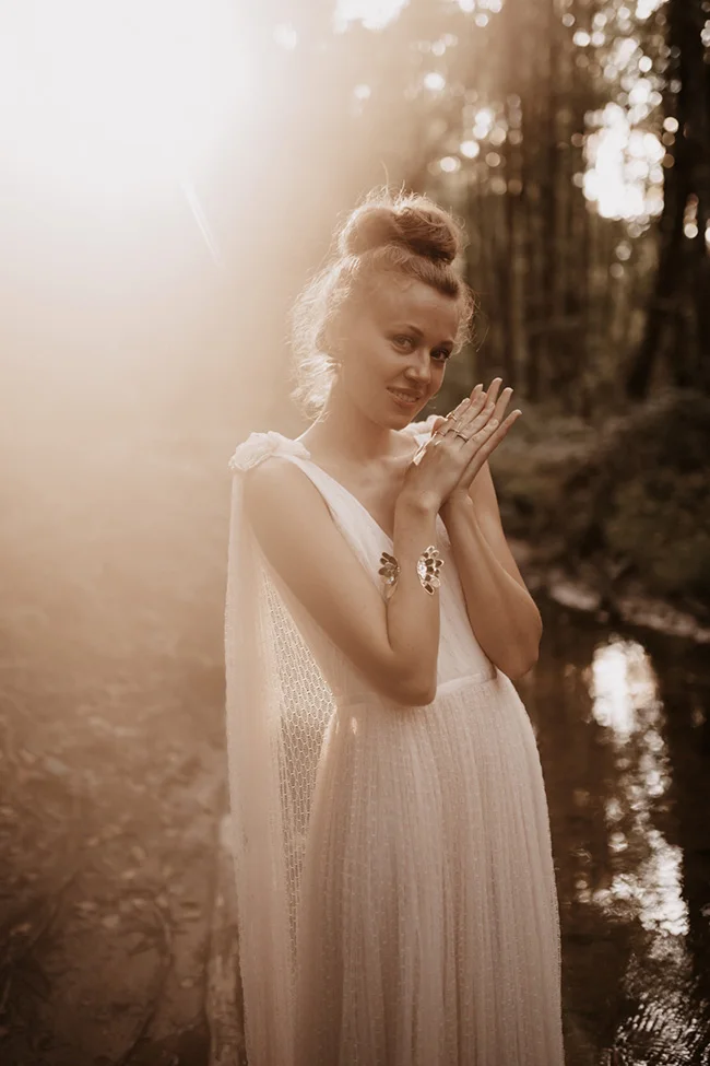 Une mariée glamour en robe blanche vaporeuse posant dans une forêt lumineuse aux tons sépia et romantiques.