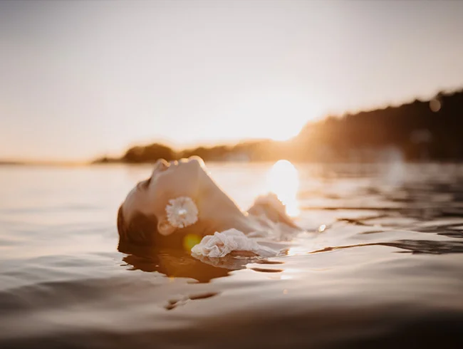 Une mariée flottant dans l'eau au coucher du soleil, une fleur blanche à l'oreille, dans une ambiance douce et onirique.
