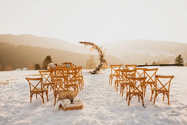 Chaises en bois disposées pour une cérémonie de mariage dans un paysage montagneux enneigé sous une lumière dorée.