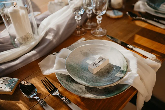 Détail d'une table de mariage élégante avec vaisselle artisanale bleue, linge blanc et bougie sur bois naturel.