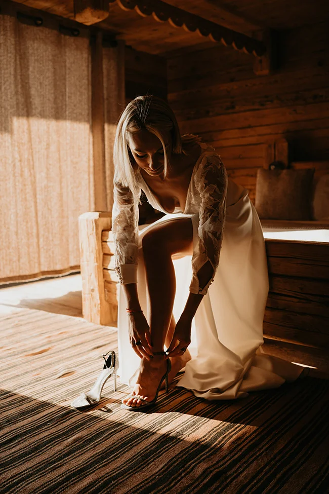 Une mariée blonde en robe blanche enfile ses talons hauts dans un chalet en bois chaleureux et lumineux.