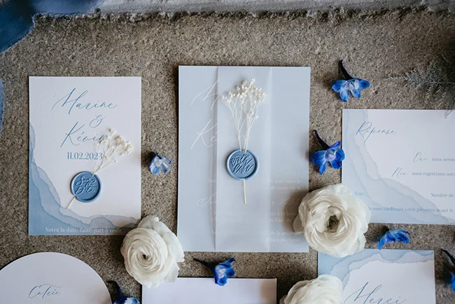 Papeterie de mariage bleue et blanche avec sceaux de cire et fleurs blanches sur un fond neutre et minéral.