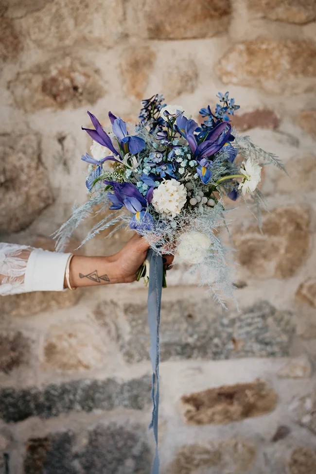 Main tenant un bouquet de fleurs bleues et blanches avec un ruban gris devant un mur de pierres rustiques.