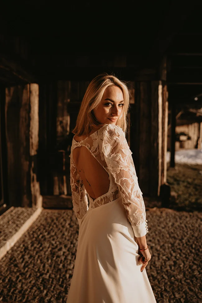 Mariée élégante en robe blanche à dentelle, posant dans un chalet rustique sous une lumière chaude et naturelle.
