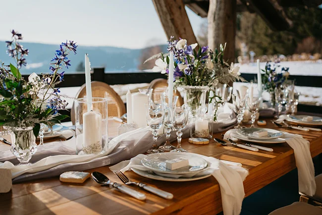 Table de mariage élégante avec fleurs blanches et bleues, bougies et nappage blanc sur fond de paysage montagneux.
