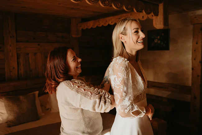 Une mariée rayonnante en robe blanche et une femme rient ensemble dans un chalet en bois chaleureux.
