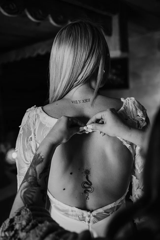 Photo noir et blanc d'une mariée de dos, aux cheveux blonds et tatouages, aidée à enfiler sa robe en dentelle.