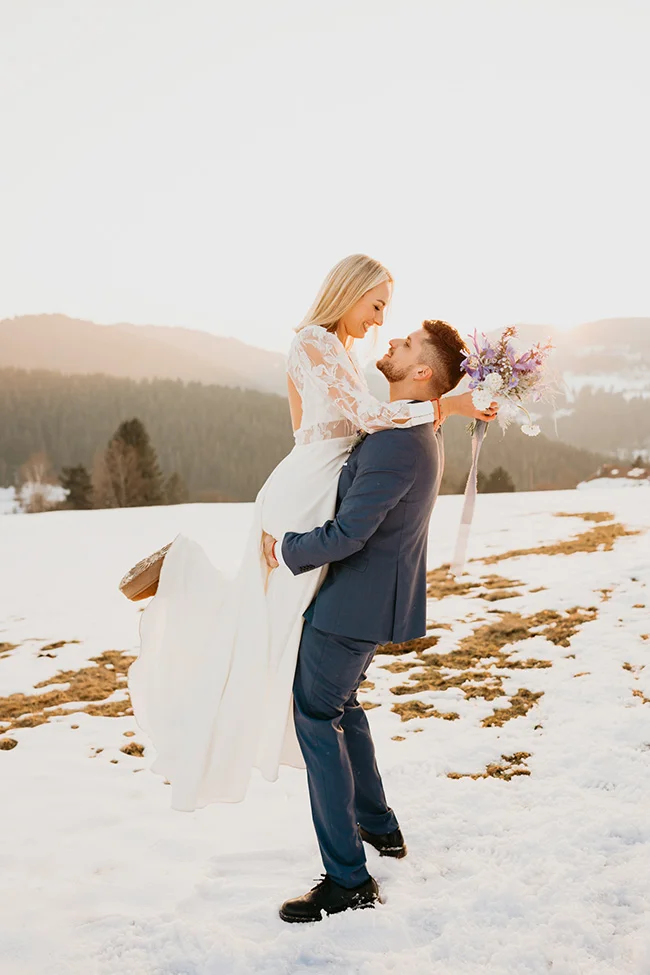 Un couple de mariés s'embrasse tendrement dans la neige, entouré de montagnes sous une lumière douce et hivernale.