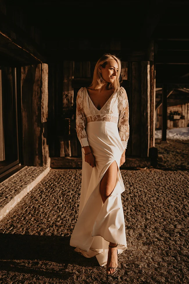 Femme en robe de mariée blanche élégante avec fente et manches en dentelle, posant devant un chalet en bois rustique.
