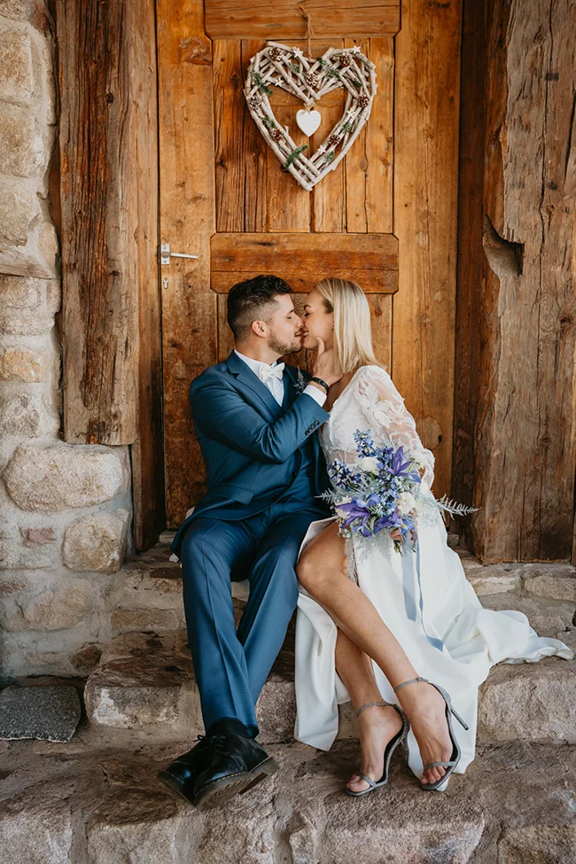 Jeunes mariés s'embrassant tendrement devant une porte en bois rustique, ambiance romantique et élégante à la montagne.