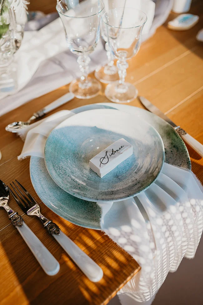 Dressage de table élégant avec assiettes bleu aquarelle, verres en cristal et marque-place sur nappe blanche.