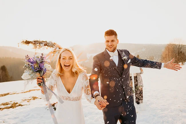 Jeunes mariés riant main dans la main dans un paysage enneigé et lumineux, avec un bouquet aux tons violets.