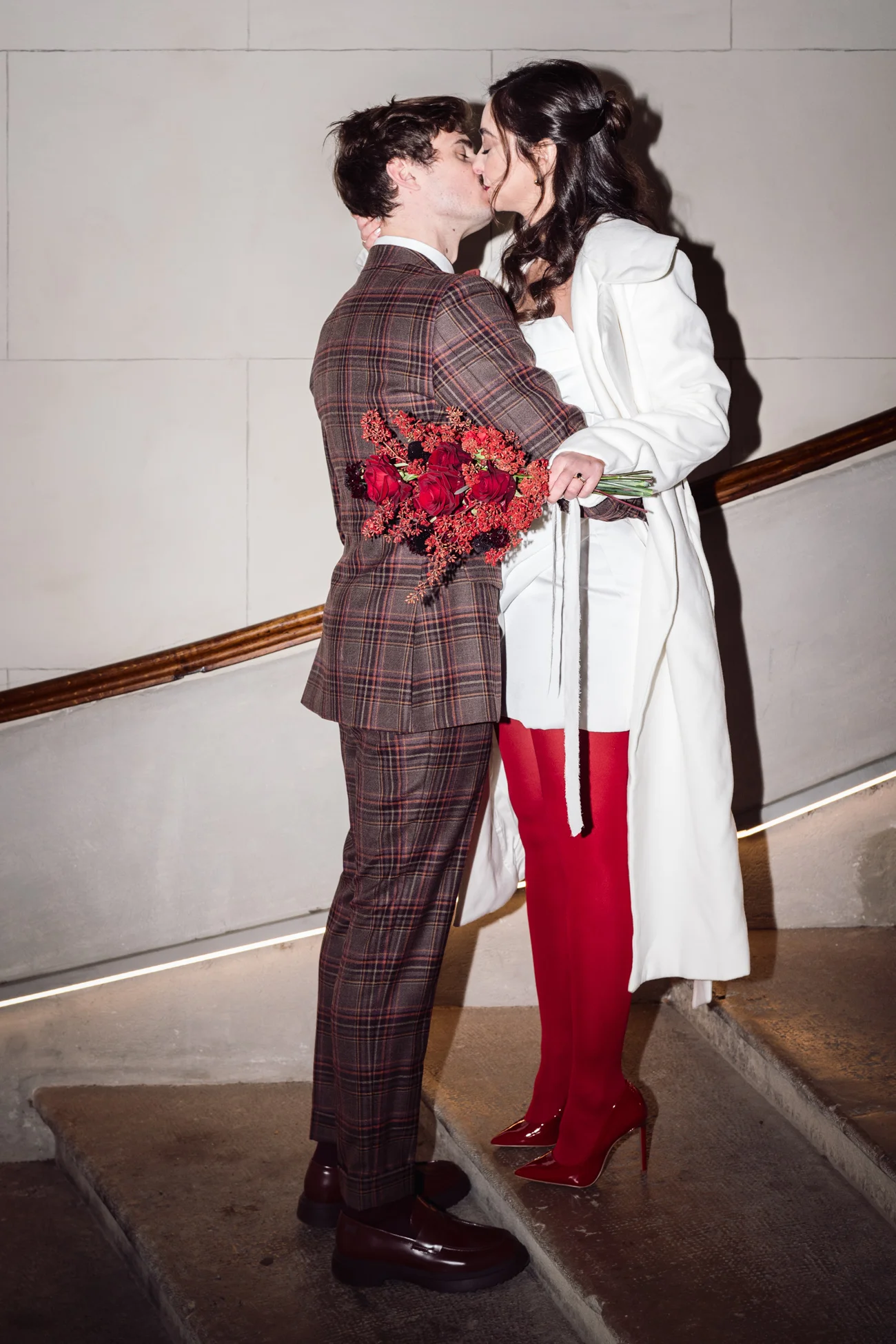 Couple s'embrassant sur un escalier, elle en manteau blanc et bottes rouges, lui en costume à carreaux rétro.