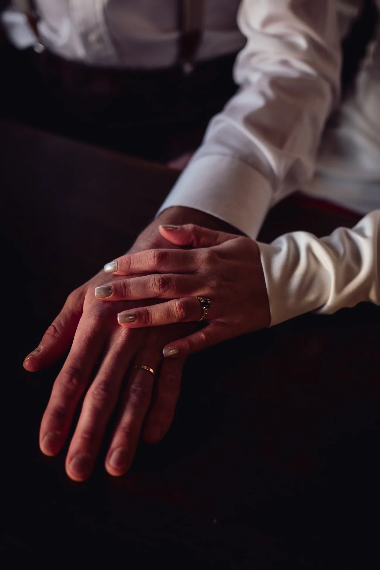 Gros plan romantique de deux mains entrelacées avec alliances sur une table sombre, ambiance rétro et chic.