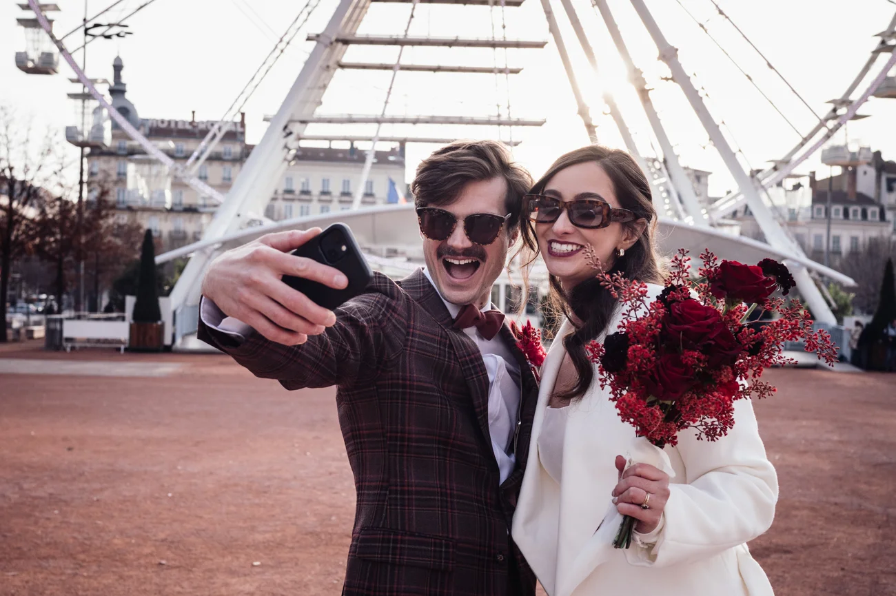 Couple rétro chic prenant un selfie avec un bouquet de roses rouges devant une grande roue urbaine.