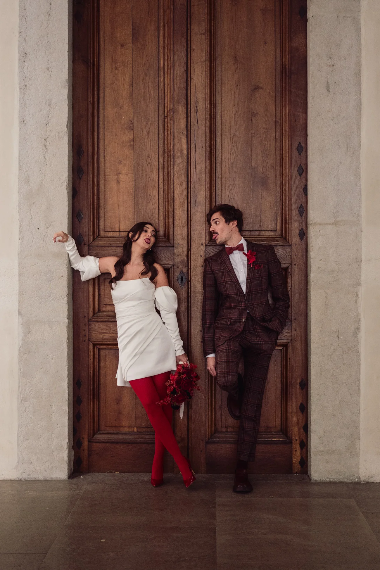 Couple rétro chic devant une porte en bois, elle en robe blanche et collants rouges, lui en costume marron à carreaux.