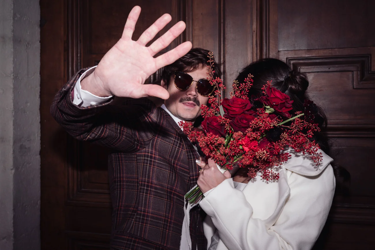 Couple rétro chic : un homme en costume à carreaux et lunettes noires et une femme en blanc cachée par des roses rouges.