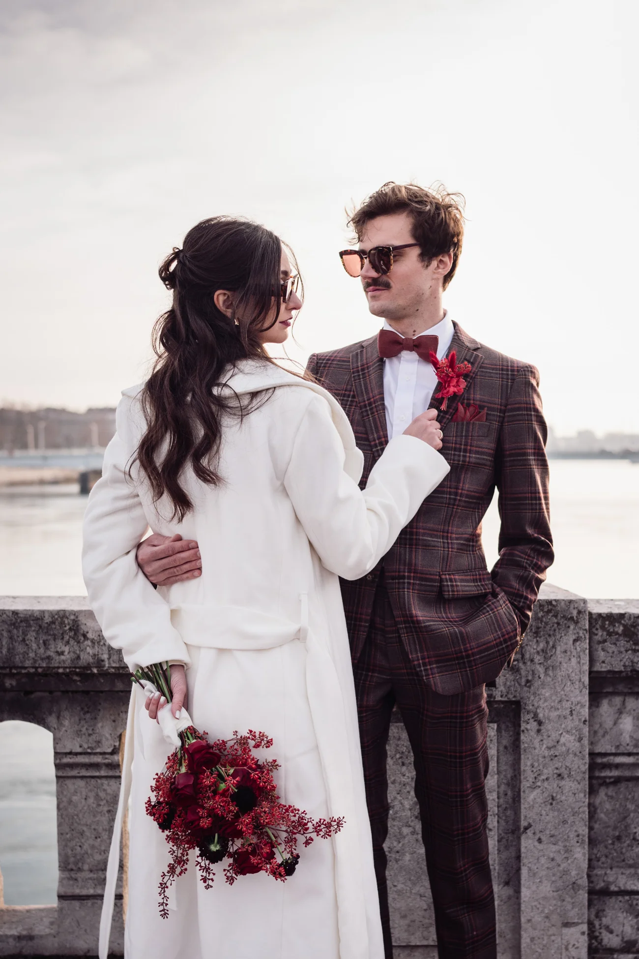 Couple rétro chic en manteau blanc et costume à carreaux marron, se regardant avec tendresse près d'un pont.