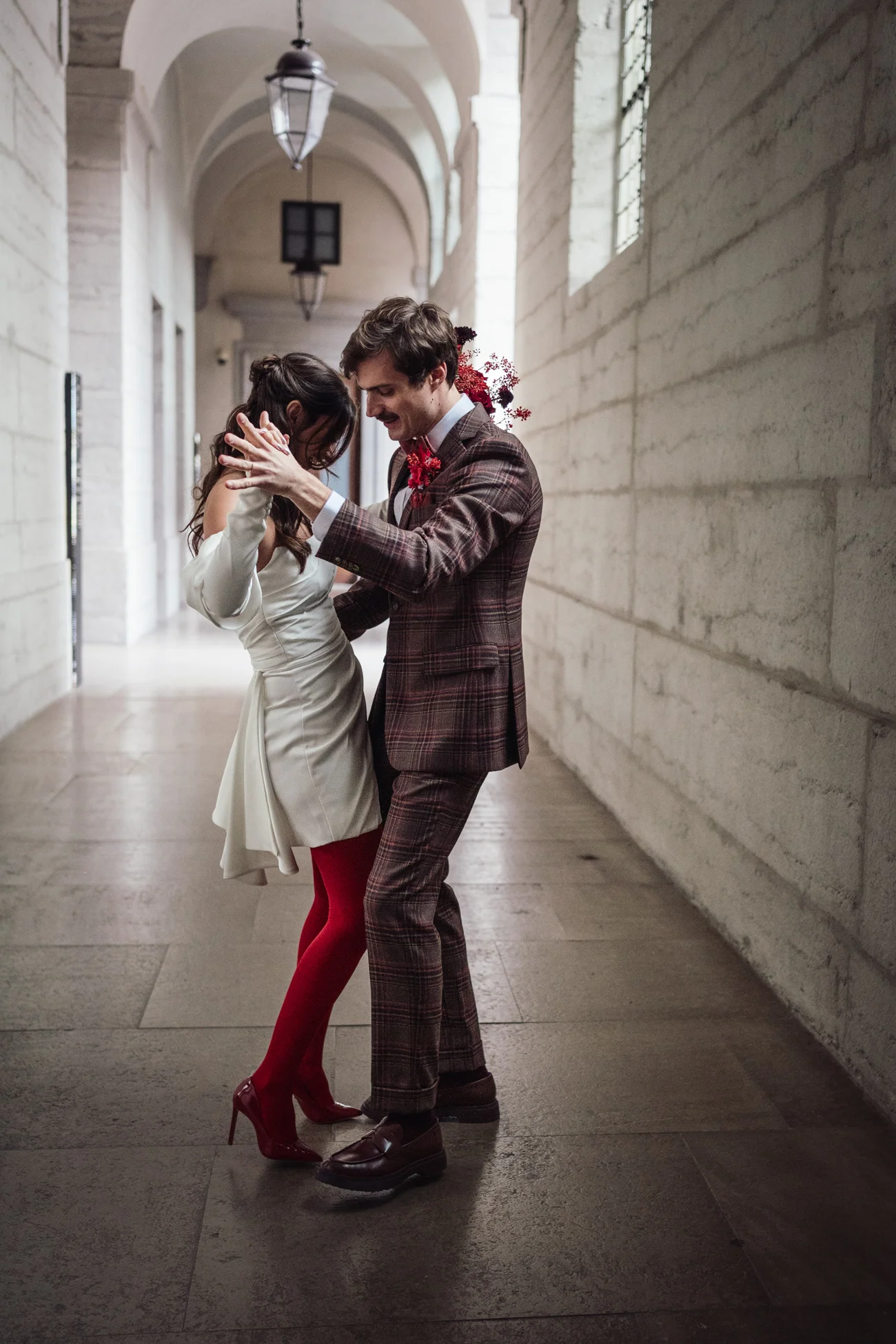 Couple en tenue rétro chic dansant dans un couloir en pierre, elle en robe blanche et collants rouges, lui en costume carreaux.