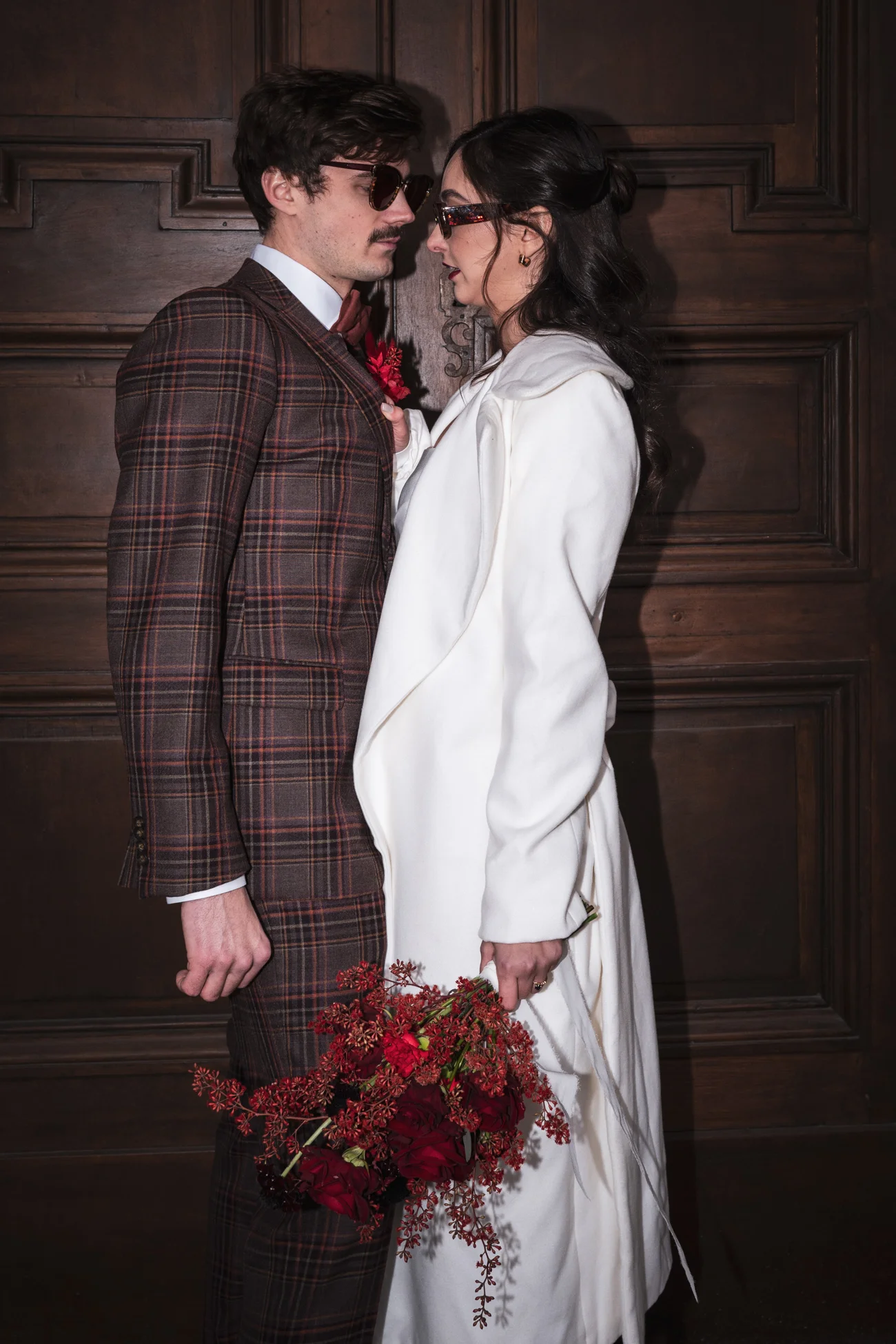 Couple rétro chic face à face devant une porte en bois, portant un costume à carreaux et une robe blanche avec bouquet rouge.