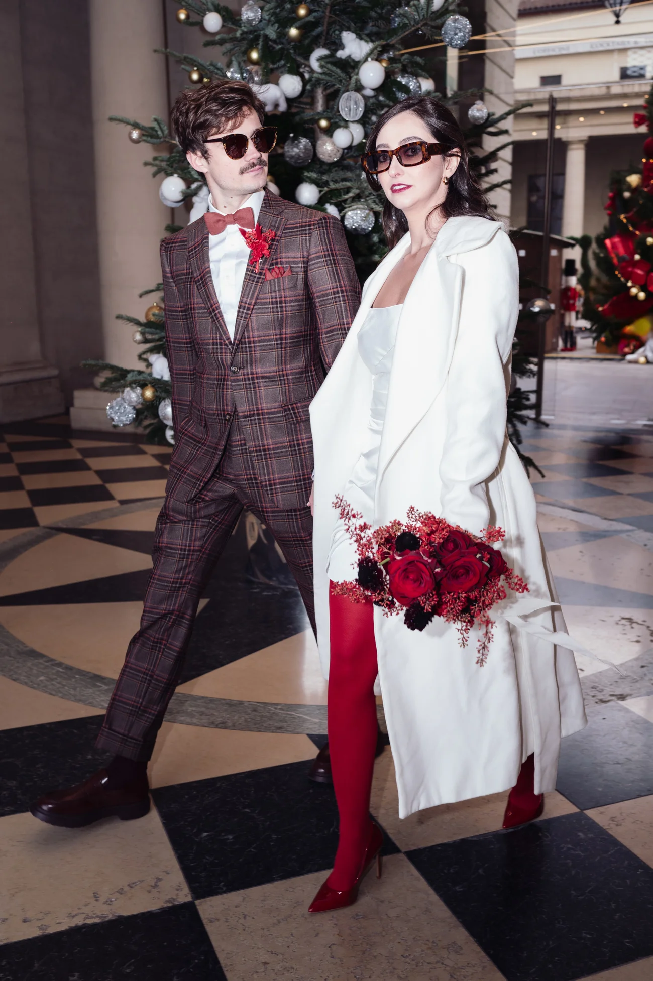 Couple rétro chic posant devant un sapin : homme en costume à carreaux et femme en manteau blanc et collants rouges.