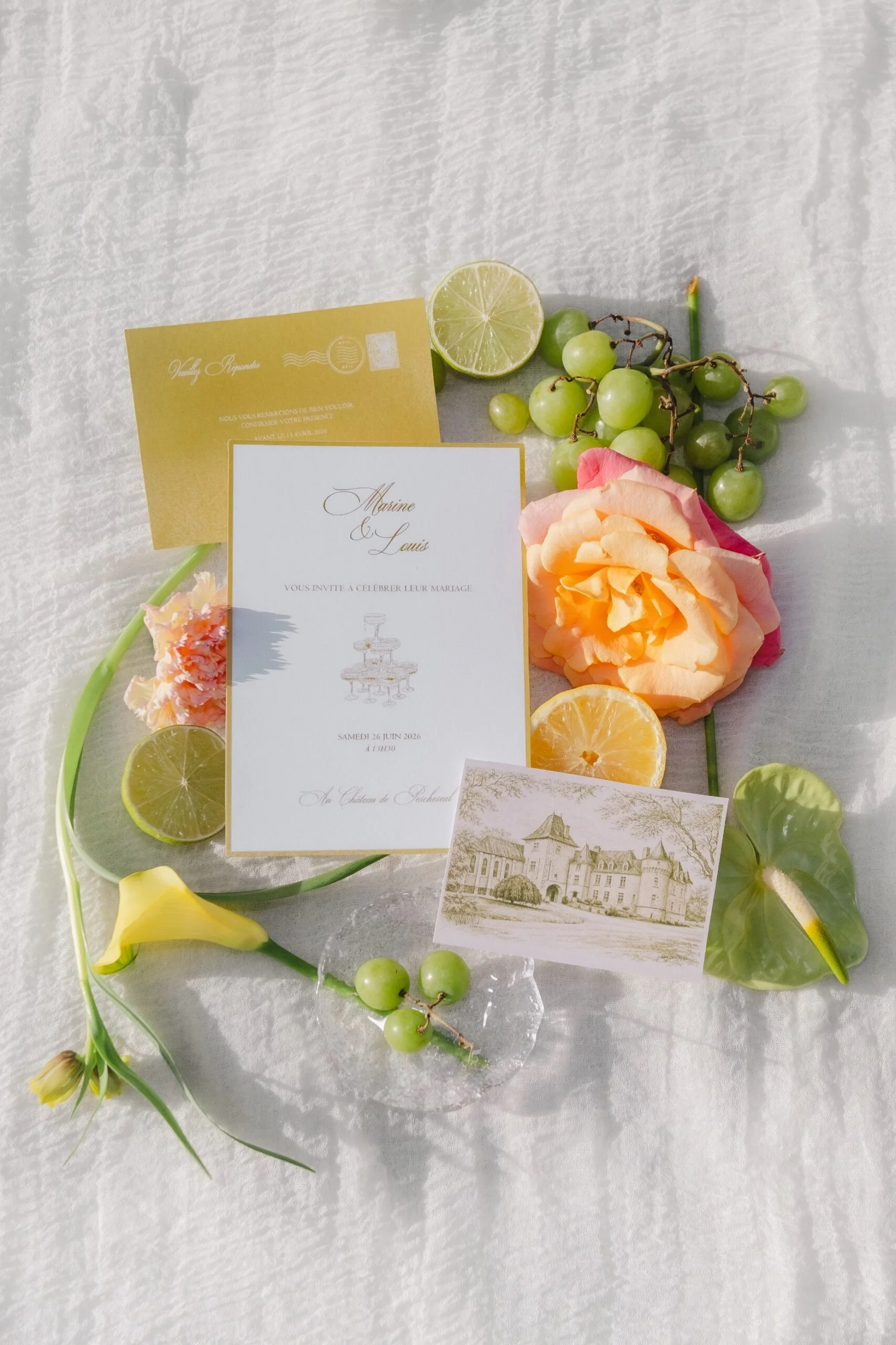 Mise en scène lumineuse de faire-part de mariage entourés de fleurs pastel, de raisins verts et de tranches de citron lime.