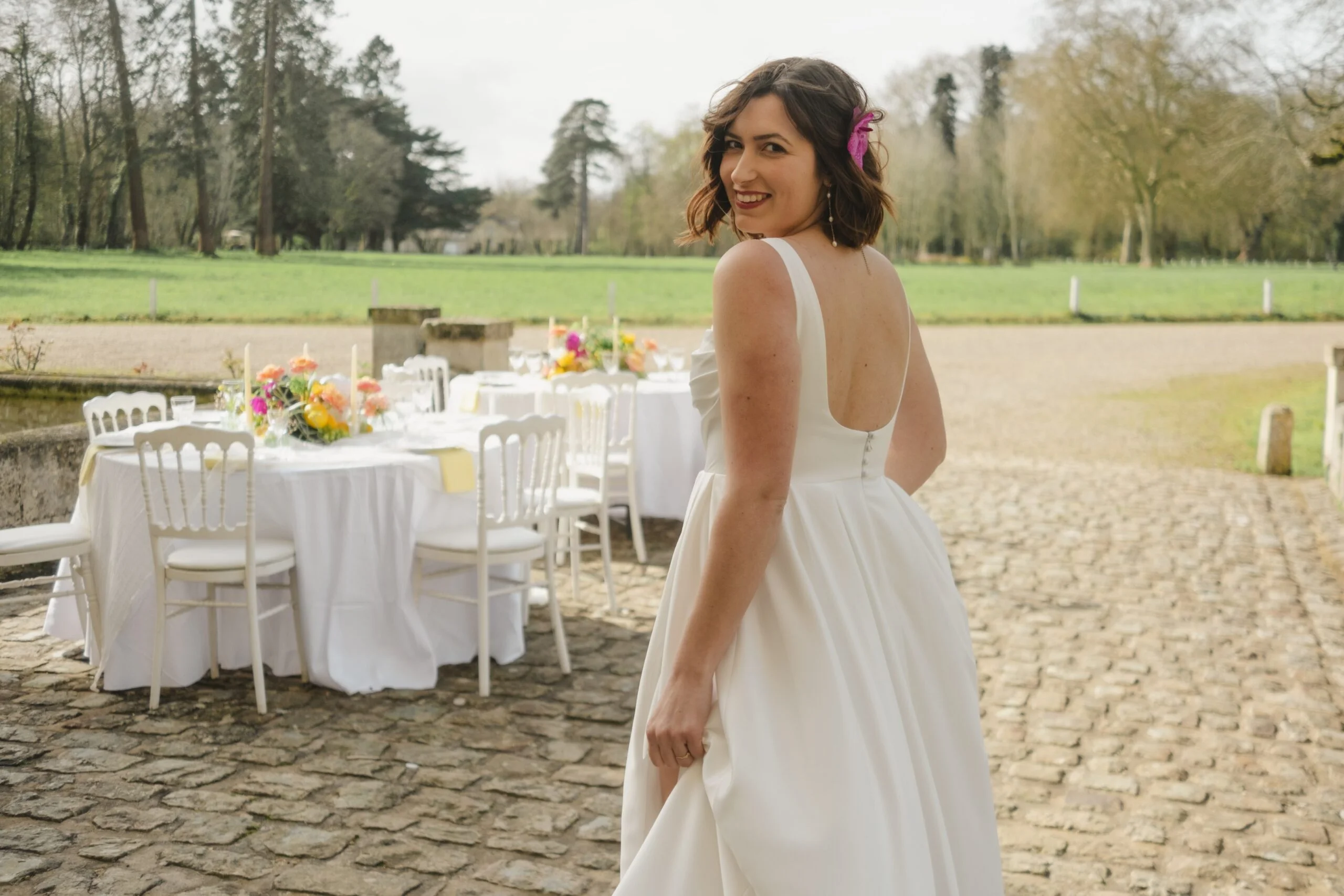 Mariée souriante en robe blanche devant des tables de réception décorées de fleurs colorées en extérieur.