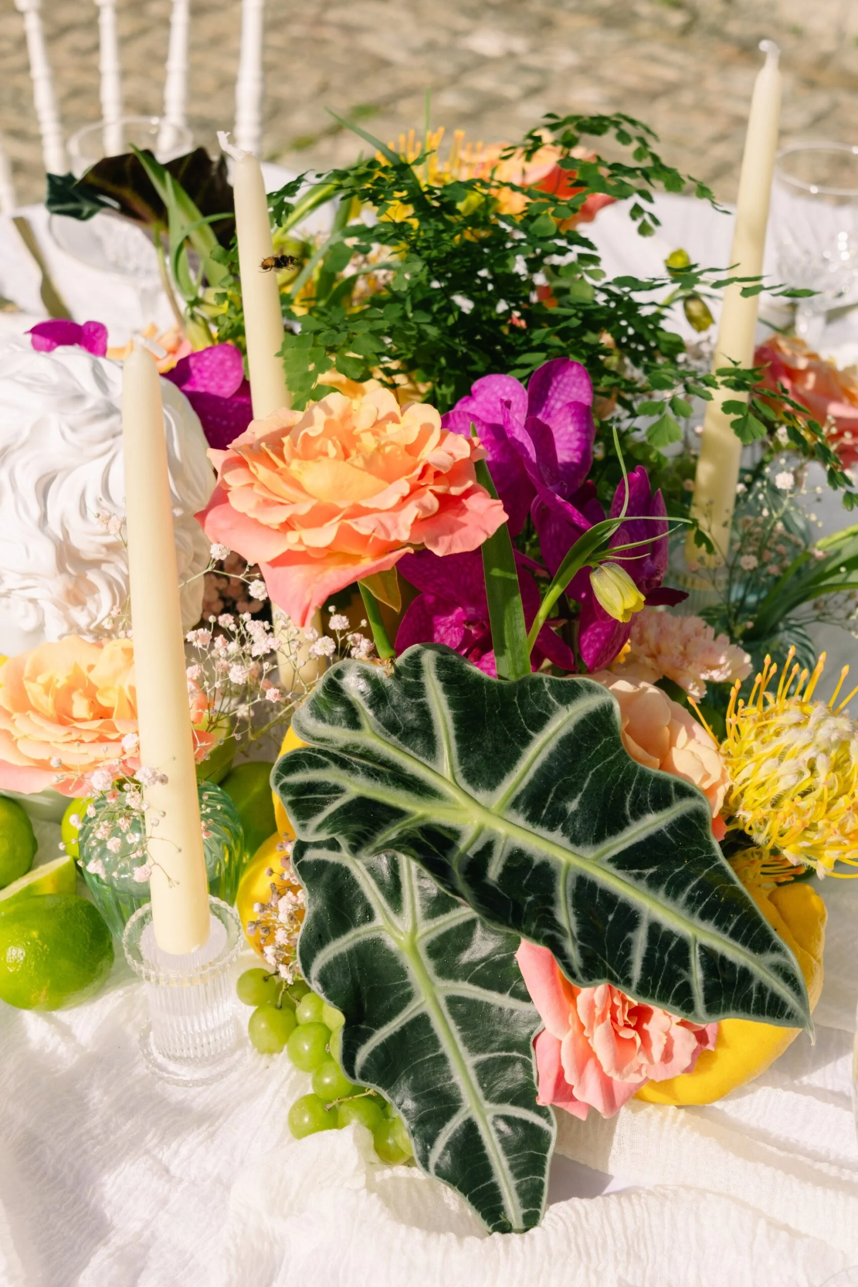 Décoration de table festive avec fleurs colorées, agrumes, raisins et bougies sur un nappage blanc.
