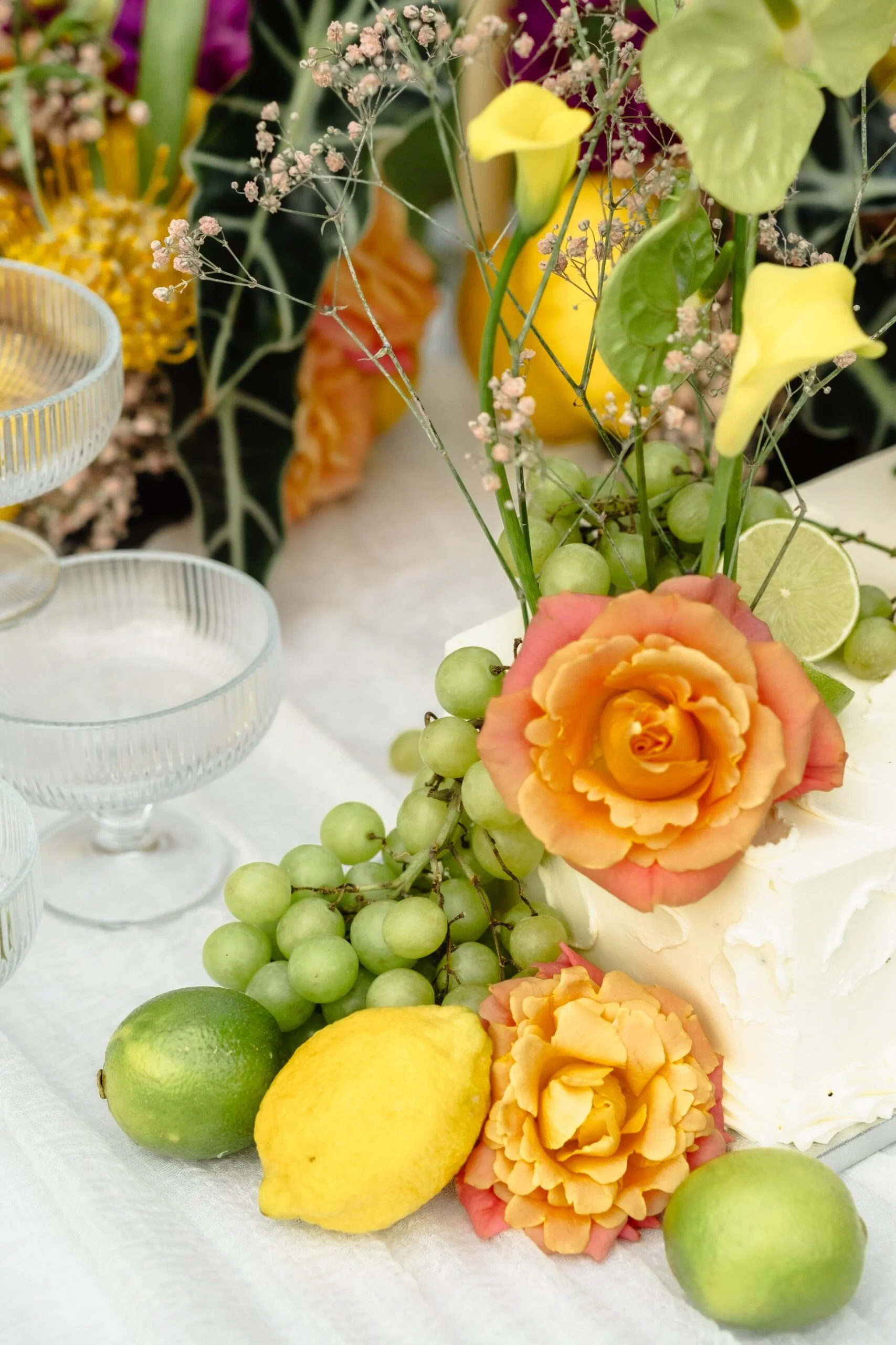 Décoration de table festive avec roses orangées, raisins verts, citrons et citrons verts sur un nappage blanc.