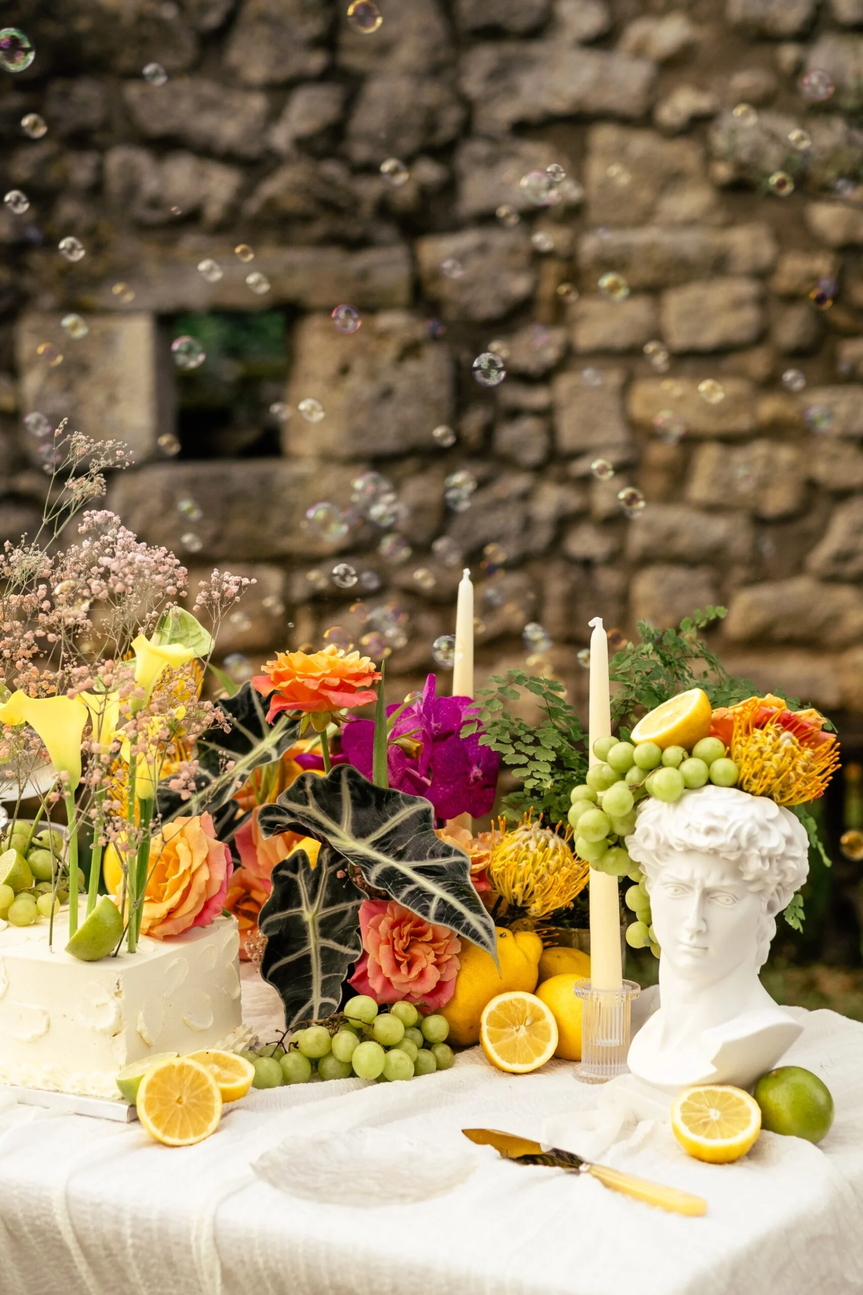 Décoration de table festive avec gâteau blanc, fleurs colorées, citrons, raisins et buste antique sous des bulles.