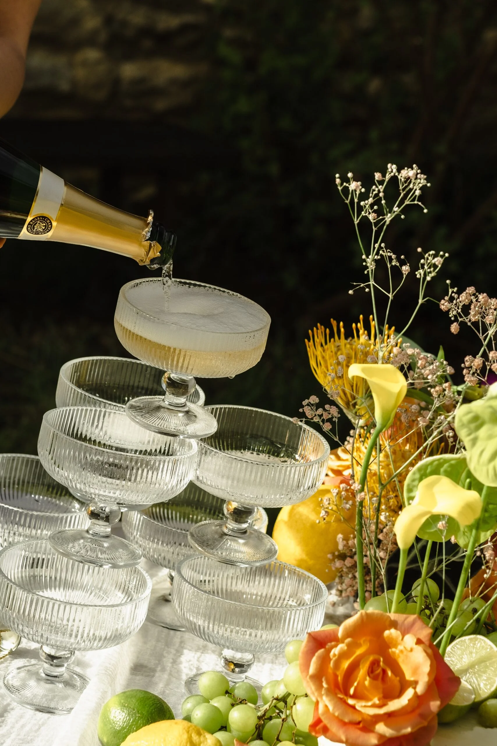 Champagne versé dans des coupes en verre entourées de fleurs colorées, d'agrumes et de raisins sur une nappe blanche.