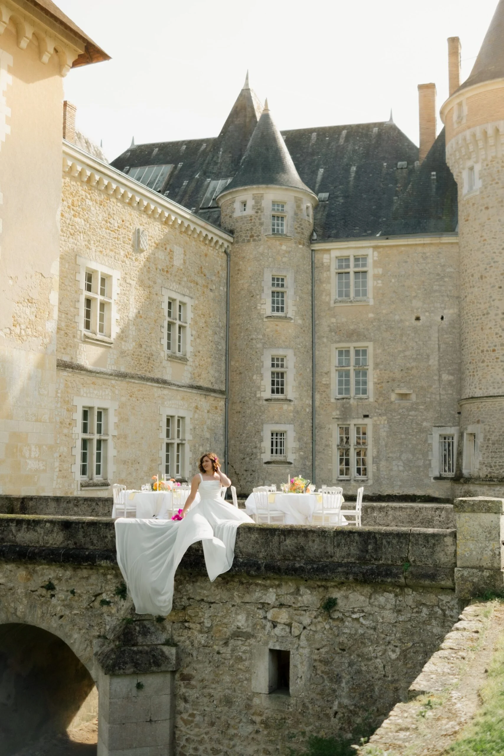 Mariée en robe blanche assise à une table décorée de fleurs sur la terrasse d'un château en pierre sous un soleil doux.