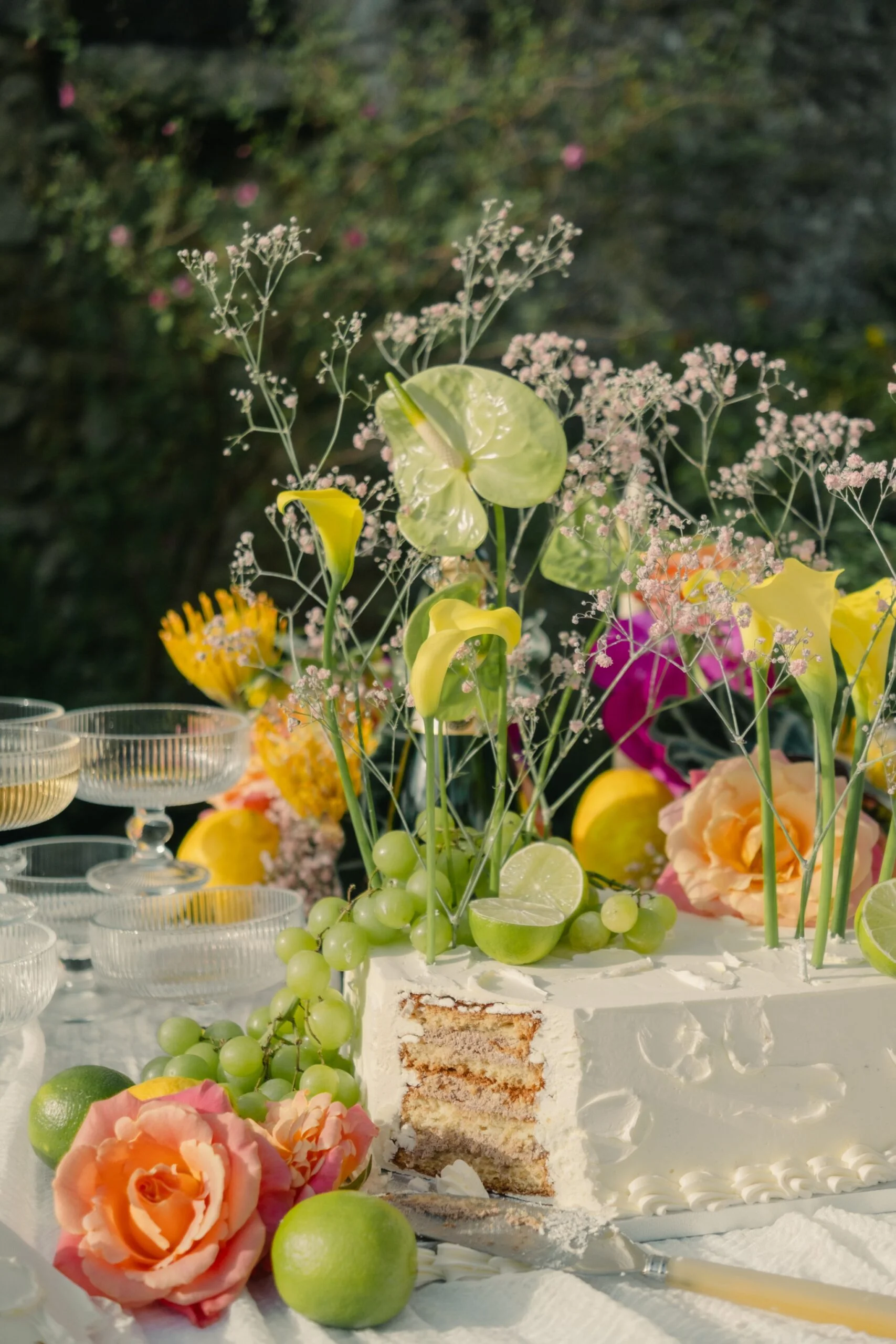 Décoration de table festive avec un gâteau blanc, des roses, des lys, des raisins et des agrumes aux tons pastel.