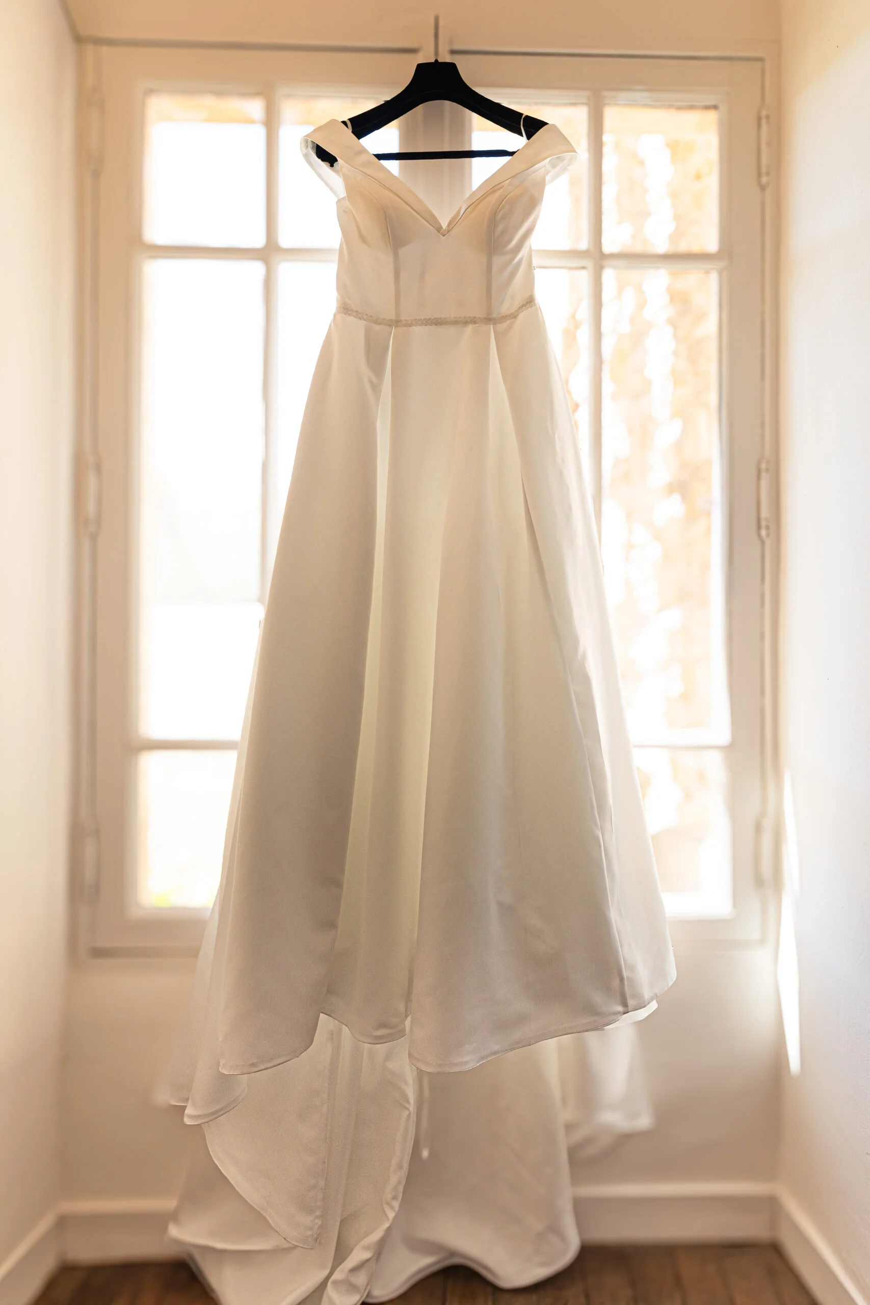 Robe de mariée blanche élégante suspendue devant une fenêtre lumineuse dans un cadre classique.
