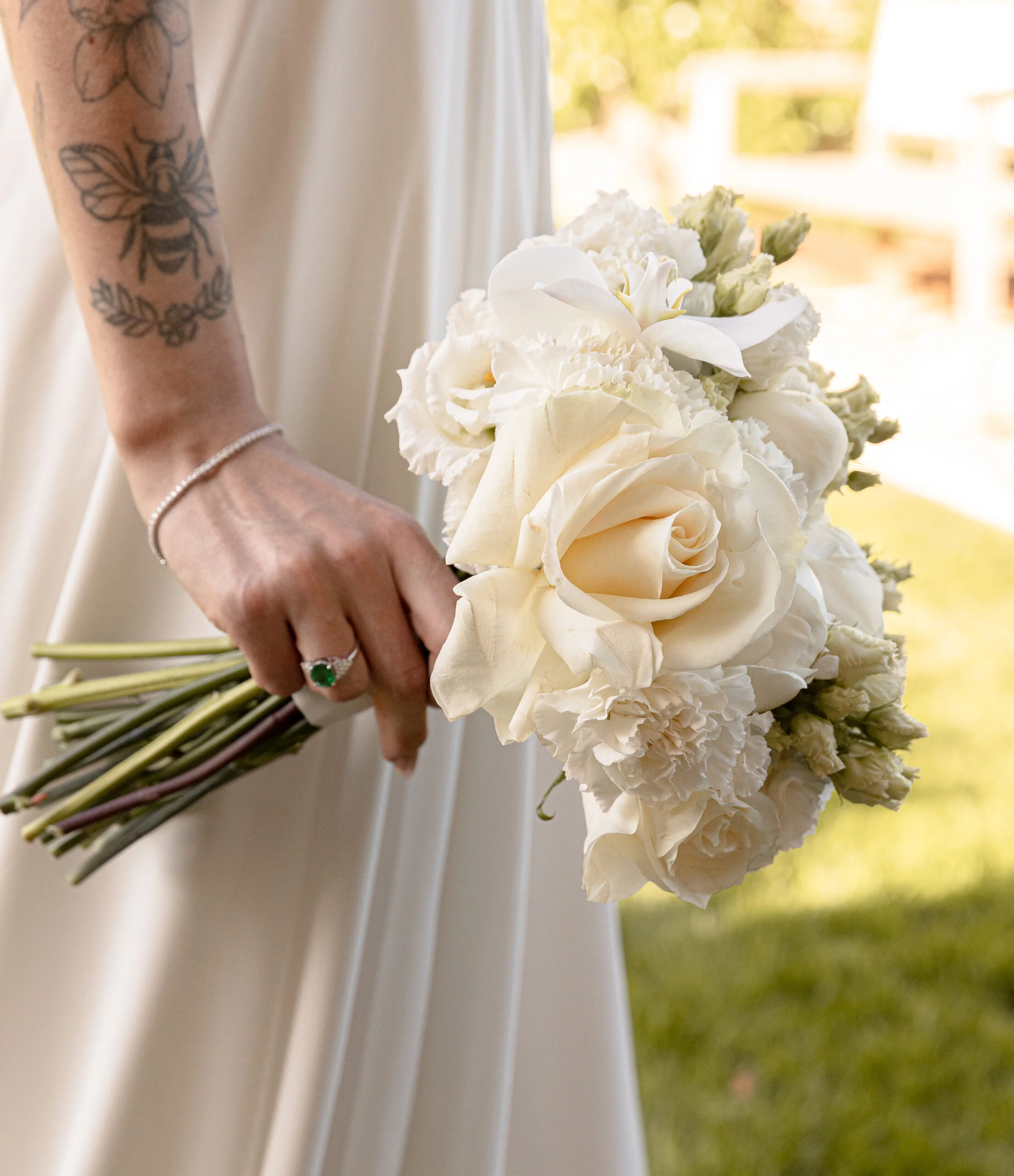 Mariée en robe blanche tenant un bouquet de roses et fleurs blanches, avec un tatouage d'abeille sur l'avant-bras.