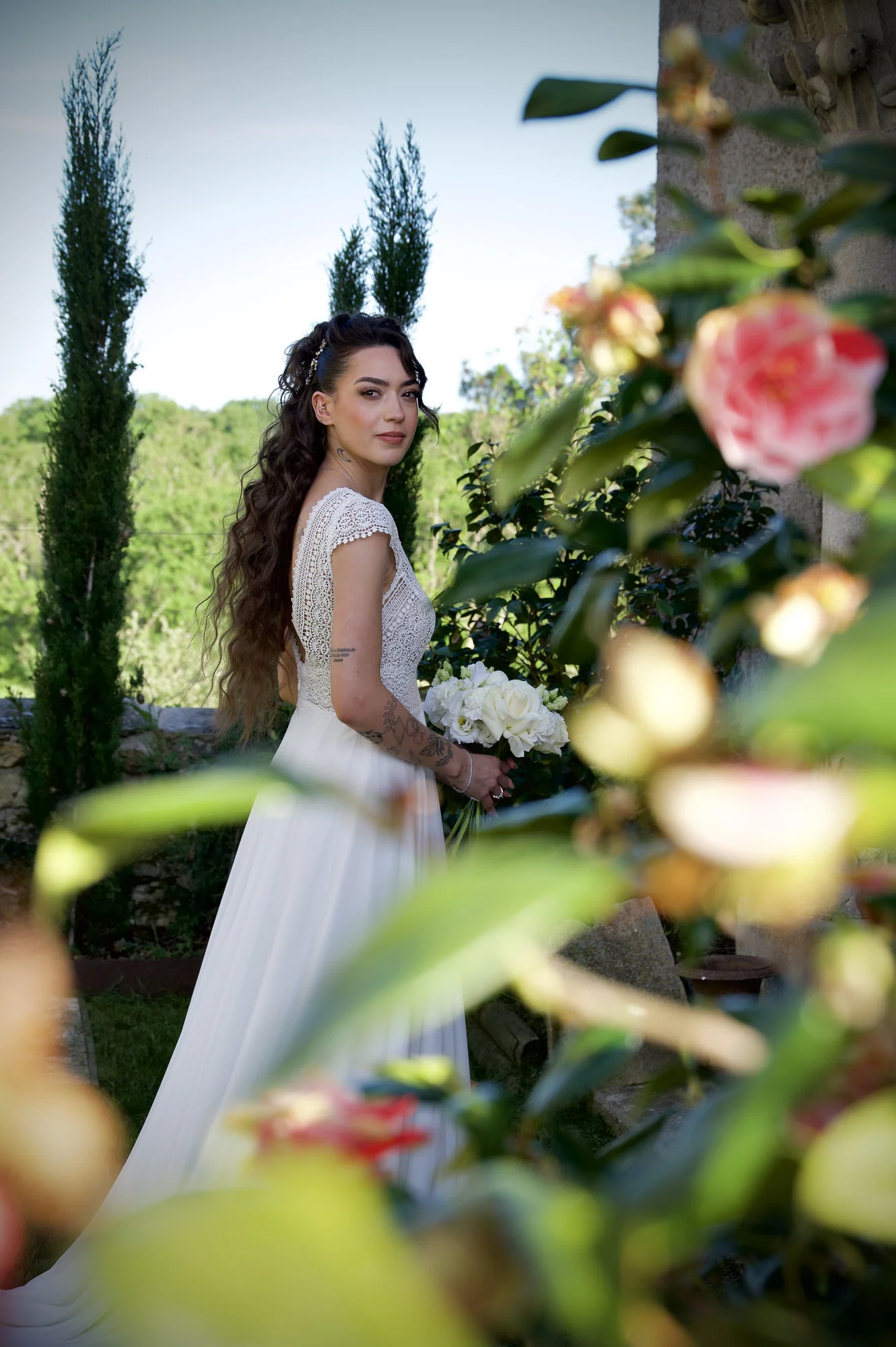 Mariée élégante en robe blanche et dentelle tenant un bouquet, posant dans un jardin luxuriant au style romantique.
