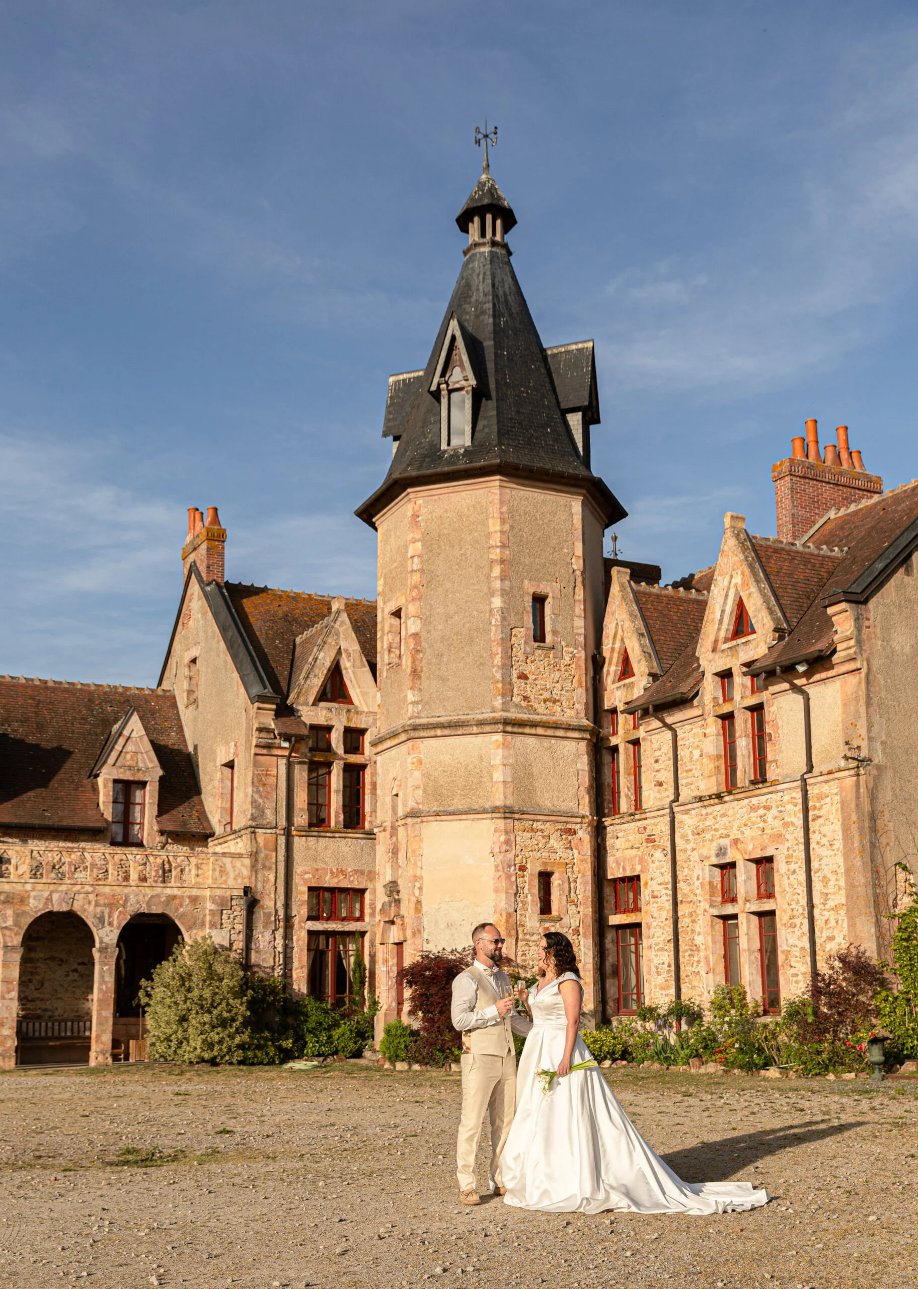 Jeunes mariés élégants posant devant un château en pierre aux tons chauds sous un ciel bleu lumineux.