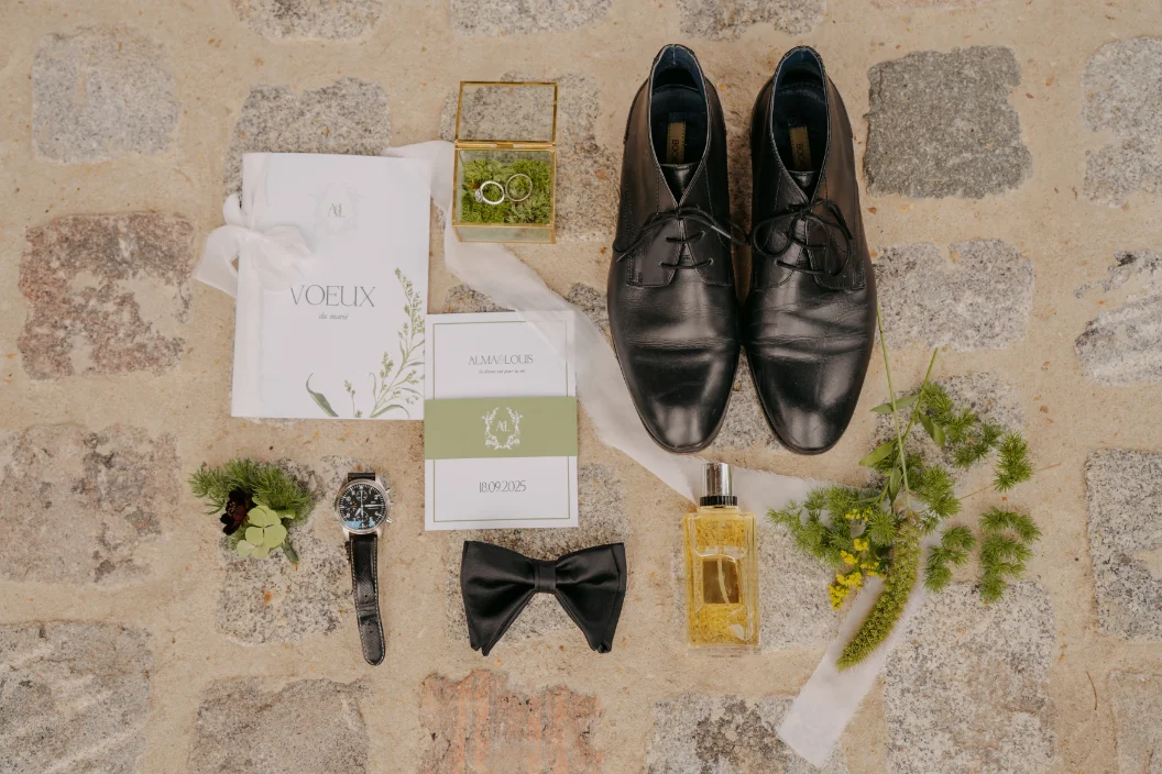 Accessoires de marié posés sur un sol en pierre : chaussures noires, nœud papillon, montre, parfum et alliances.