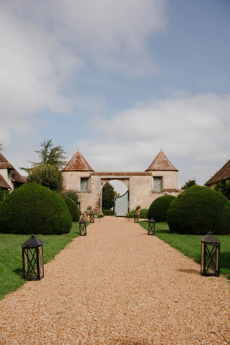 Allée de gravier menant à une porte cochère ancienne encadrée de tourelles et de buis taillés sous un ciel clair.