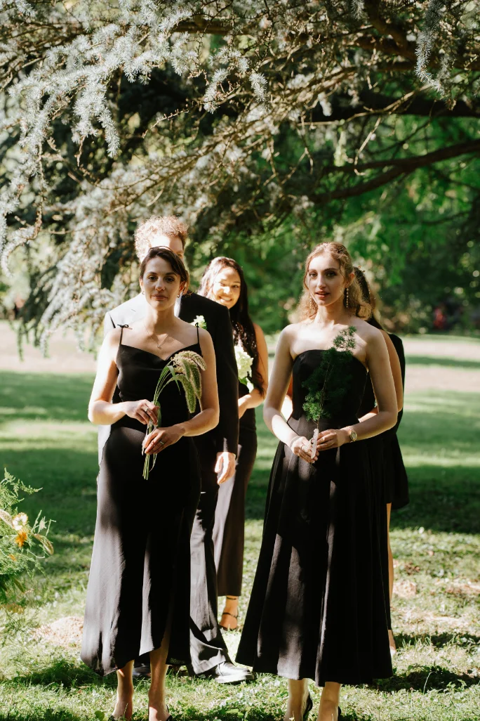 Quatre personnes élégantes en tenues noires posant sous un arbre fleuri dans un jardin lumineux.