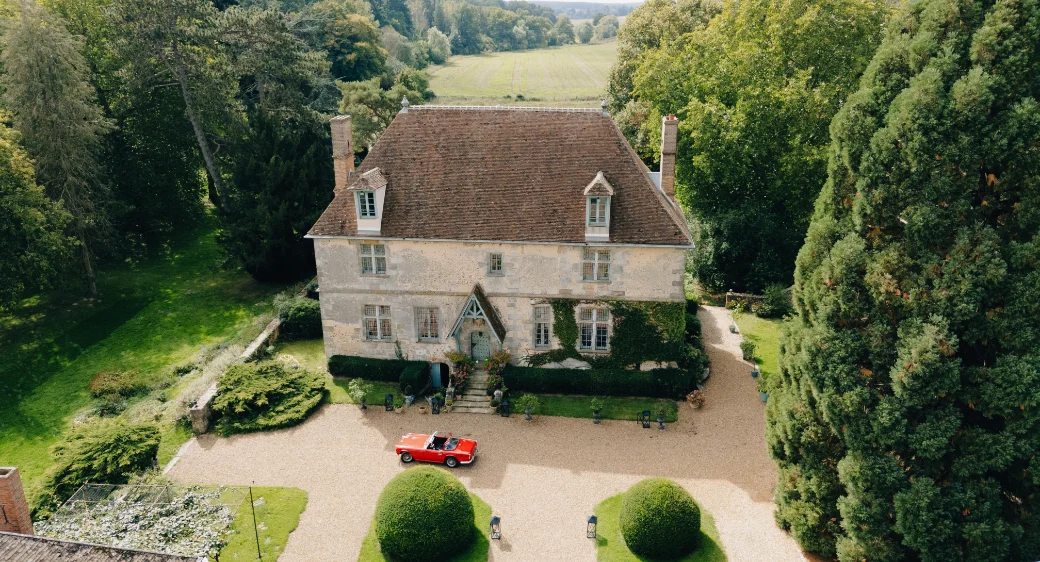 Manoir ancien en pierre avec une voiture décapotable rouge garée devant, entouré d'un jardin verdoyant et luxuriant.