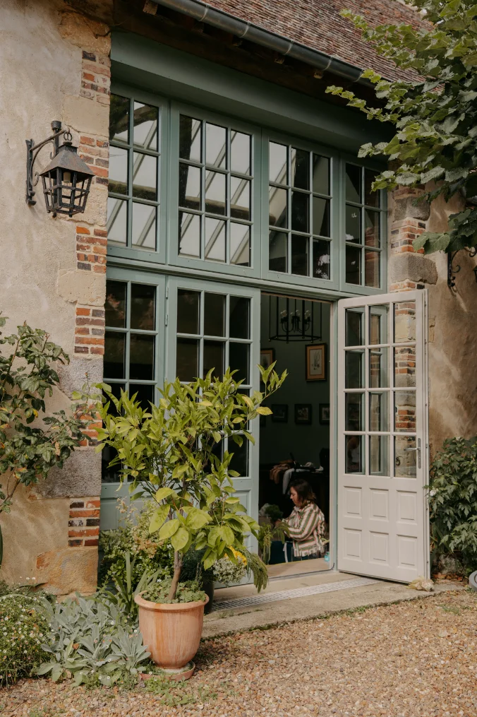 Entrée d'une maison de campagne aux menuiseries vert sauge, avec une plante en pot et une femme à l'intérieur.